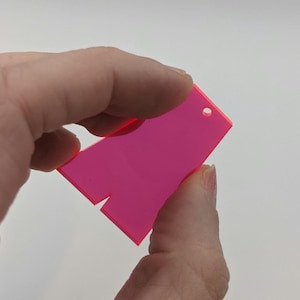 Könnte beinhalten: Ein neonrosa Acrylschild mit einem schlüssellochförmigen oberen Teil und einer Kerbe am unteren Rand. Das Schild wird zwischen zwei Fingern vor einem weißen Hintergrund gehalten. Das Schild ist ein leuchtendes Pink.