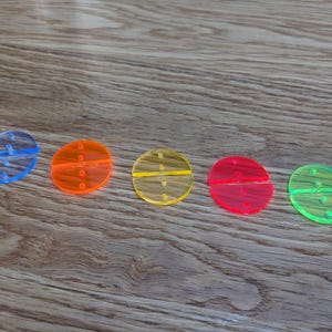 Könnte beinhalten: Fünf farbenfrohe, ovale Spielsteine aus Acryl in Blau, Orange, Gelb, Rot und Grün. Jedes Teil hat einen horizontalen Schlitz und drei kleine Löcher. Die Teile sind auf einer Holzoberfläche angeordnet.