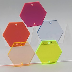 Könnte beinhalten: Eine Sammlung farbenfroher, sechseckiger Acryl-Anhänger in Pink, Orange, Gelb, Grün und Blau. Jeder Anhänger hat ein Loch zum Auffädeln oder Befestigen. Die Anhänger sind in einem geschichteten, überlappenden Muster angeordnet.