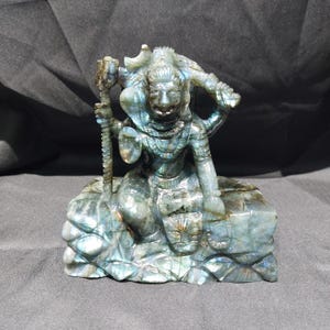 May include: A carved stone sculpture of a deity, featuring a seated figure with multiple arms and a serene expression. The sculpture is predominantly blue-green with iridescent flashes, and it rests on a textured base. The figure holds a staff.