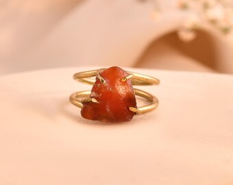 May include: A gold-colored ring featuring a raw, reddish-brown gemstone. The ring has a double-band design, with the stone held in place by small prongs. The gemstone has a natural, irregular shape, adding to its unique appeal. The ring is set against a soft, neutral background.
