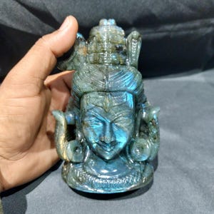 May include: A carved labradorite sculpture of a deity with intricate details. The sculpture features a serene face, ornate headdress, and serpent motifs. The stone displays iridescent blue and green hues, creating a captivating visual effect. The sculpture is held by a hand.