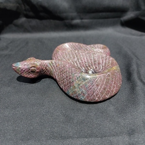 Może przedstawiać: Rzeźba węża z rzeźbionego kamienia w odcieniach czerwieni, fioletu i zieleni. Wąż jest zwinięty, z szczegółowymi wytrawionymi wzorami na całym ciele. Rzeźba jest umieszczona na ciemnym tle.