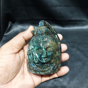 May include: A carved gemstone sculpture of a serene face with intricate details. The sculpture is held in a hand, showcasing its blue and green hues. The carving has a loop at the top, suggesting it could be a pendant or ornament.