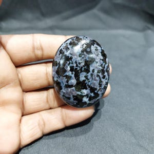 May include: An oval gemstone with a speckled pattern of black and blue-grey. The polished stone is held in a hand, showcasing its smooth surface and natural variations. The gemstone is a unique piece of jewellery.