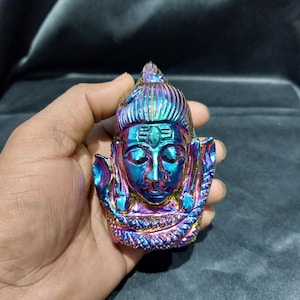 May include: A colorful, iridescent sculpture of a deity's head. The sculpture features shades of blue, purple, and gold. The head has detailed facial features and is held in a hand.
