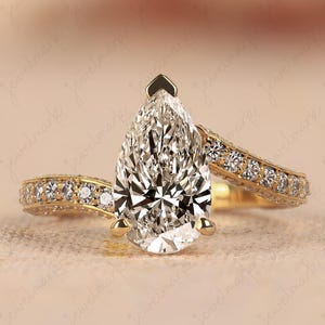 May include: A gold engagement ring with a pear-shaped diamond center stone. The band features small, sparkling stones and a delicate design. The ring is set against a soft, neutral background.