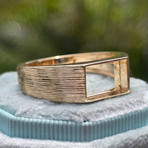 May include: A gold-colored ring with a rectangular setting. The band has a textured, ridged design. The ring is displayed on a light blue velvet surface.