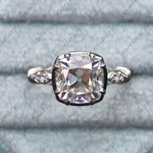May include: A silver-toned ring featuring a large, cushion-cut center stone held by four prongs. The band is crafted with a split-shank design, and each side is accented with three small, round, clear stones set into the metal shoulders.