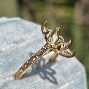 May include: A gold-colored ring setting with a floral design. The ring features a detailed band with an engraved pattern and a prong setting designed to hold a gemstone. The ring is displayed on a light blue surface.