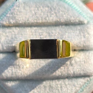 May include: A gold ring with a rectangular black onyx stone. The ring has a polished gold band and a simple, elegant design. The ring is displayed on a light blue surface.