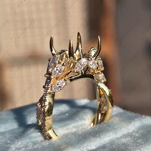 May include: A gold-colored ring with a unique design, featuring marquise-cut and round gemstones. The ring has a detailed, nature-inspired motif with a crown-like top. The ring is set on a blue velvet surface.