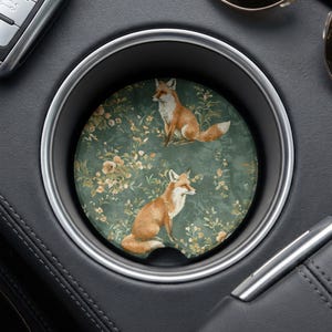 May include: A circular car cup holder coaster featuring a dark green background with a repeating pattern of orange foxes and small beige flowers. The coaster has a small semi-circular notch at the bottom edge for easy removal from the holder.