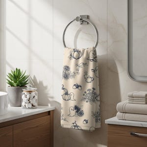 Peut inclure: Une serviette à main couleur crème suspendue à un anneau argenté, ornée d'un motif répétitif en lignes bleu foncé représentant des fantômes, citrouilles, glands, pommes de pin, fleurs et théières, formant un design textile automnal.