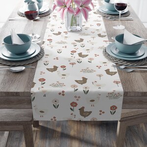 Peut inclure: Une nappe blanche avec un motif répété de poulets bruns et blancs, et de fleurs rouges et roses. La nappe est posée sur une table en bois, avec des couverts et un vase de lys roses.