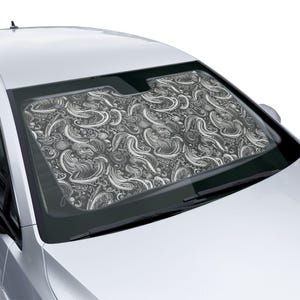 May include: A car windshield sun shade featuring a black background with a white, intricate, swirling paisley-inspired pattern. The rectangular shade is positioned inside the front windshield of a silver vehicle, covering the glass area.