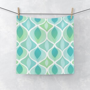 May include: A square, patterned towel with a repeating design of overlapping teardrop shapes in shades of teal, turquoise, and green, outlined in white. The towel is hanging from a clothesline with wooden clothespins.