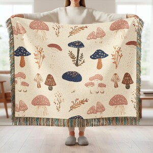 May include: A cream-colored woven throw blanket featuring a repeating pattern of various mushrooms in shades of blue, brown, and pink, interspersed with small sprigs of foliage. The blanket is finished with a multi-colored fringed edge.
