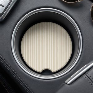 Op de afbeelding: Een ronde bekerhouder-onderzetter voor in de auto met een verticaal krijtstreepmotief in afwisselende tinten beige en crème. De onderzetter heeft een kleine halfronde inkeping aan de onderrand om het verwijderen uit de bekerhouder te vergemakkelijken.