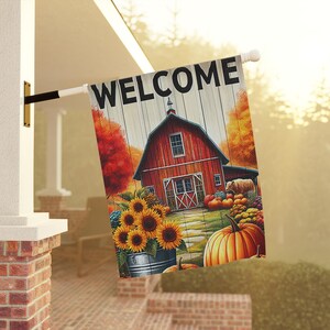 Puede incluir: Una bandera de jardín rectangular con la palabra WELCOME en letras mayúsculas negras en la parte superior. El diseño muestra un granero rojo, un cubo de metal lleno de girasoles amarillos y calabazas naranjas sobre árboles otoñales.