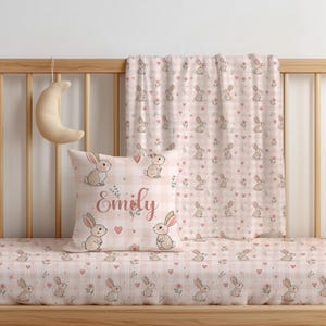 Könnte beinhalten: Ein Babybett-Set mit rosa-weißem Vichy-Muster, verziert mit Hasen, Herzen und Blumen. Das Set beinhaltet ein Spannbettlaken, ein Kissen mit dem Namen "Emily" und eine passende Decke. Ein Halbmond hängt an dem Kinderbett.