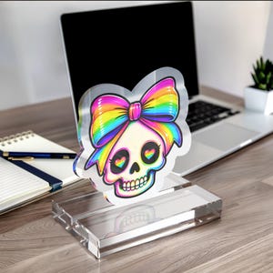 May include: A clear acrylic desk sign featuring a colorful illustration of a skull with a large rainbow-striped bow on its head and heart-shaped pupils in its eyes. The sign is mounted on a rectangular, transparent acrylic base stand.