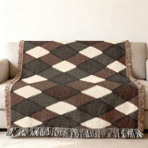 Op de afbeelding: Een geweven plaid met een herhalend ruitpatroon in tinten crème, donkerbruin en antracietgrijs. De deken heeft een getextureerd uiterlijk met zichtbare draaddetails en een decoratieve franje langs de boven- en onderranden.