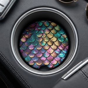 May include: A circular car cup holder coaster featuring a vibrant, iridescent mermaid scale pattern in shades of teal, purple, gold, and blue. The coaster has a small semi-circular notch at the bottom edge and fits inside a silver-rimmed holder.