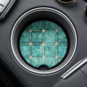 May include: A circular car cup holder coaster featuring a repeating teal and white geometric tile pattern. The coaster has a small semi-circular notch at the bottom edge and is positioned inside a black vehicle cup holder with a silver trim ring.