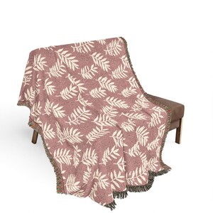 May include: A woven throw blanket featuring a dusty rose background with a repeating pattern of cream-coloured fern leaves. The blanket has a textured knit construction and is finished with decorative multi-coloured fringe along the edges.