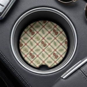 May include: A circular car cup holder coaster featuring a beige, green, and red plaid pattern. The design is decorated with repeating illustrations of brown pinecones and green pine needles arranged diagonally across the surface of the coaster.