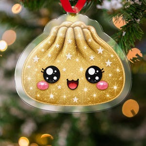 May include: A clear acrylic ornament shaped like a dumpling with a gold glitter finish and white star accents. The dumpling features a printed face with large black eyes, pink cheeks, and a smiling mouth. A red ribbon is attached to the top loop.