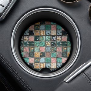 May include: A circular car cup holder coaster featuring a grid pattern of small, square tiles in shades of teal, dusty pink, and muted gold. Each tile displays a weathered, crackled texture, creating a mosaic effect within the silver-rimmed holder.