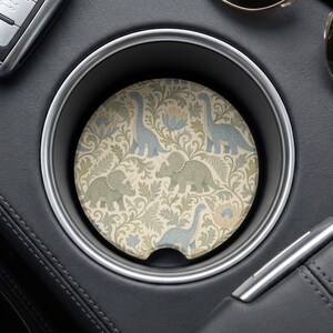 May include: A circular car cup holder coaster featuring a repeating pattern of light blue and sage green dinosaurs, including triceratops and long-necked sauropods, interspersed with beige and blue floral and leafy botanical motifs on a cream background.