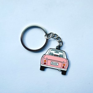 May include: A pink car keyring with the word "FIGARO" on the number plate. The keyring has a silver ring and chain. The car has a white roof and black tyres. The background is a light blue.