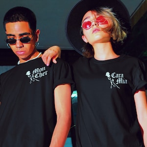 May include: Two people wearing black t-shirts with white text and a rose graphic. The text on the left shirt says "Mon Cher" and the text on the right shirt says "Cara Mia".
