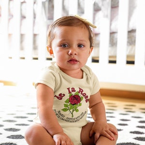May include: A baby wearing a cream-colored bodysuit with a red rose graphic and the text "De la Rosa. Fragile - Handle with care.  With De Cacaoahute Estilo Mazapan."