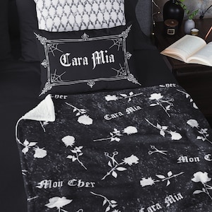 May include: A black and white bedding set featuring a decorative pillow with the text "Cara Mia" and a matching throw blanket. The blanket has a rose and scissors pattern. A patterned pillow and black sheets complete the set.