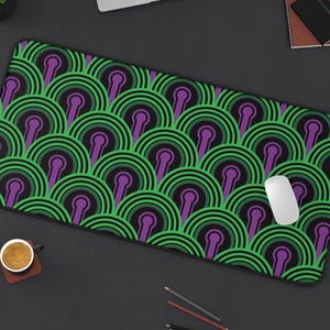 May include: A rectangular desk mat featuring a repeating geometric pattern of concentric circles in shades of green, purple, and black. The mat has a smooth surface and black edges, providing a large area for computer peripherals and office accessories.