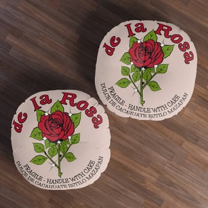 Op de afbeelding: Twee ronde, beige kussens met een rode roos en de tekst "de la Rosa" en "FRAGILE - HANDLE WITH CARE". Ze liggen op een bruin houten oppervlak.