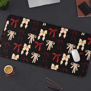 May include: Black rectangular desk mat featuring a pattern of red and cream bows. A white computer mouse rests on the mat, alongside a laptop, a small plant, and stationery. The mat is designed to protect the desk surface, measuring approximately 80 cm in length.