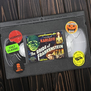 Puede incluir: Una caja de cinta VHS con fondo negro y una imagen colorida de Boris Karloff en la película "La novia de Frankenstein". La caja tiene varias pegatinas, incluyendo "Éxitos de Halloween", "Terror", "Clásico de culto", "Alquiler de 2 días" y "Creemos que te divertirás rebobinando".