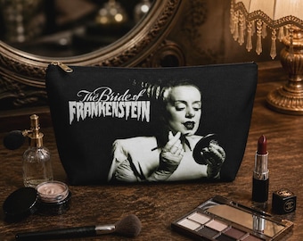 May include: Black cosmetic bag featuring an image of the Bride of Frankenstein with the text "The Bride of Frankenstein". The bag is surrounded by makeup items, including lipstick, eyeshadow, and a makeup brush.
