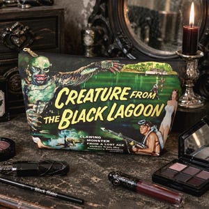 Può includere: Un beauty case nero con cerniera dorata, raffigurante un poster cinematografico vintage di "Creature from the Black Lagoon". La grafica è verde, nera e bianca. Gli articoli per il trucco sono disposti intorno alla borsa.