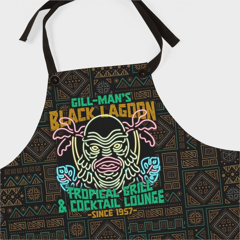 Pu&ograve; includere: Grembiule nero con un design tropicale con il testo "Gill-Man's Black Lagoon Tropical Grill & Cocktail Lounge Since 1957". Il grembiule ha uno sfondo nero con un motivo geometrico ripetuto e lacci neri.