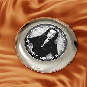 May include: A silver compact mirror with a black and white image of a woman with dark hair. The mirror has a quote around the image that reads "For the spider is chaos for the fly. Normal is an illusion. What is normal for the"