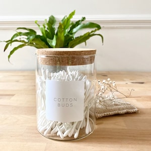 May include: A clear glass jar with a cork lid filled with white cotton buds. The jar has a white label that reads "COTTON BUDS".