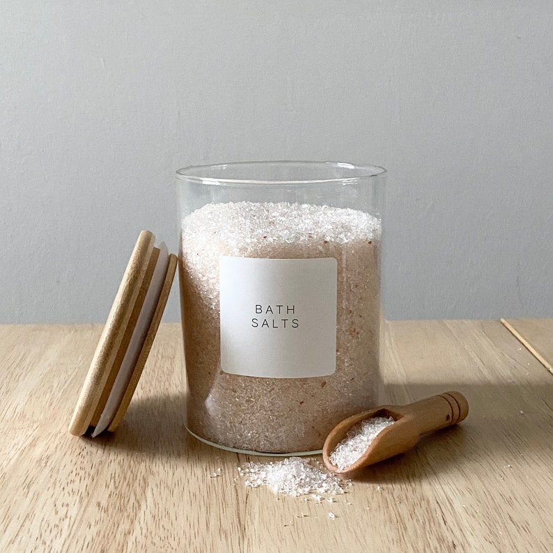 May include: A glass jar filled with pink bath salts with a white label that reads "Bath Salts". The jar has a wooden lid and a wooden scoop is next to the jar.