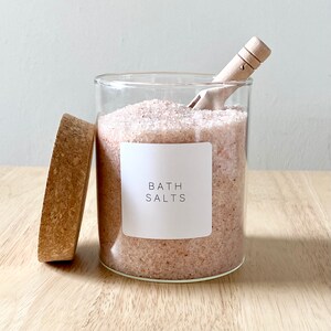 May include: A glass jar filled with pink bath salts, with a wooden scoop and a cork lid. The jar has a white label that says "Bath Salts".