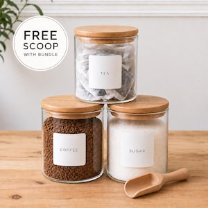 May include: Three clear glass canisters with light wood lids, each featuring a white square label with black text reading TEA, COFFEE, or SUGAR. The canisters contain tea bags, coffee granules, and white sugar. A small wooden scoop rests in front.